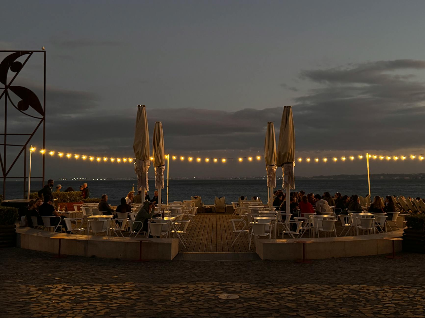 Outdoor dining terrace in Lisbon at twilight with string lights and harbor view