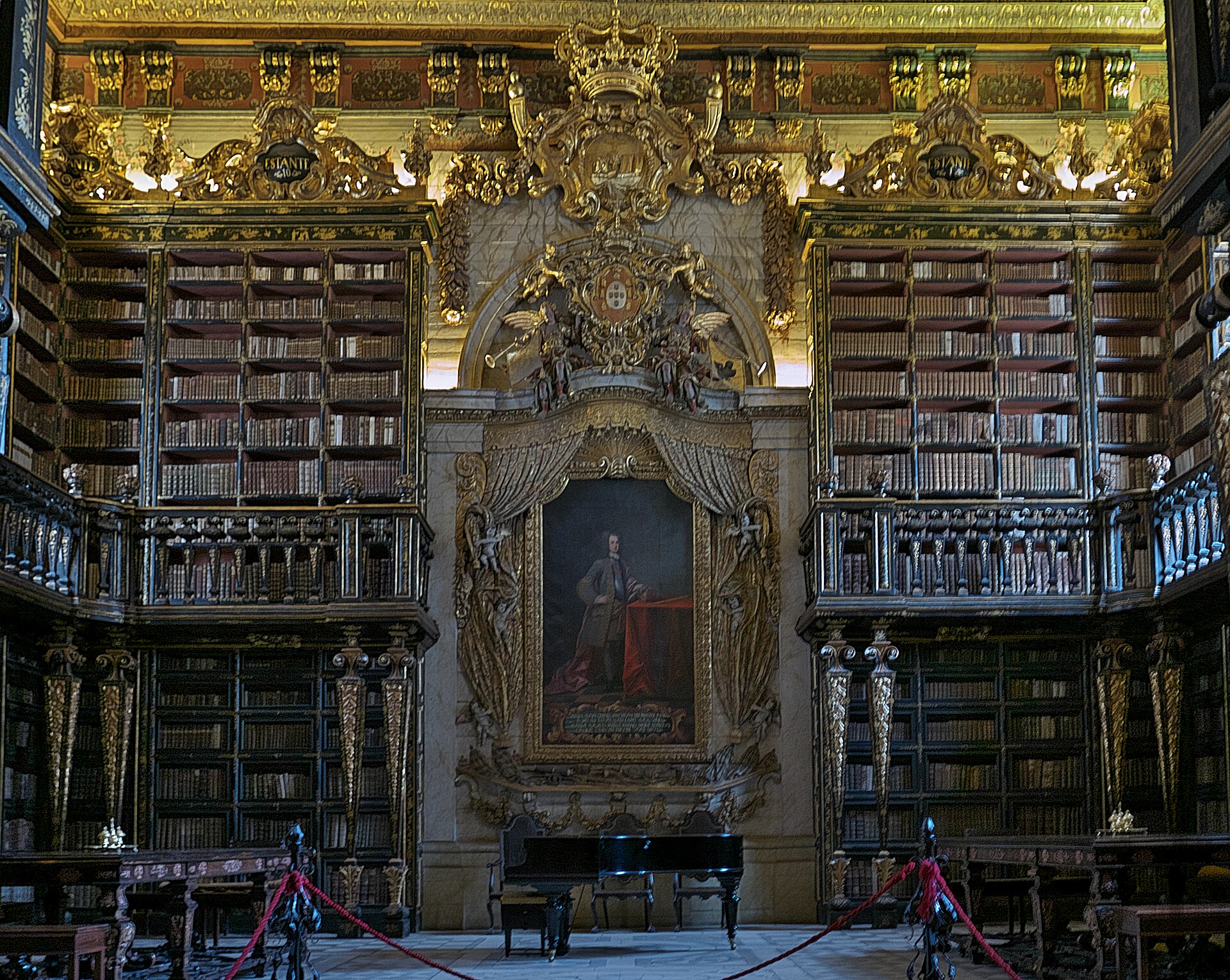 The gilded baroque interior of the Biblioteca Joanina at the University of Coimbra, with tiered bookshelves and a portrait of King João V