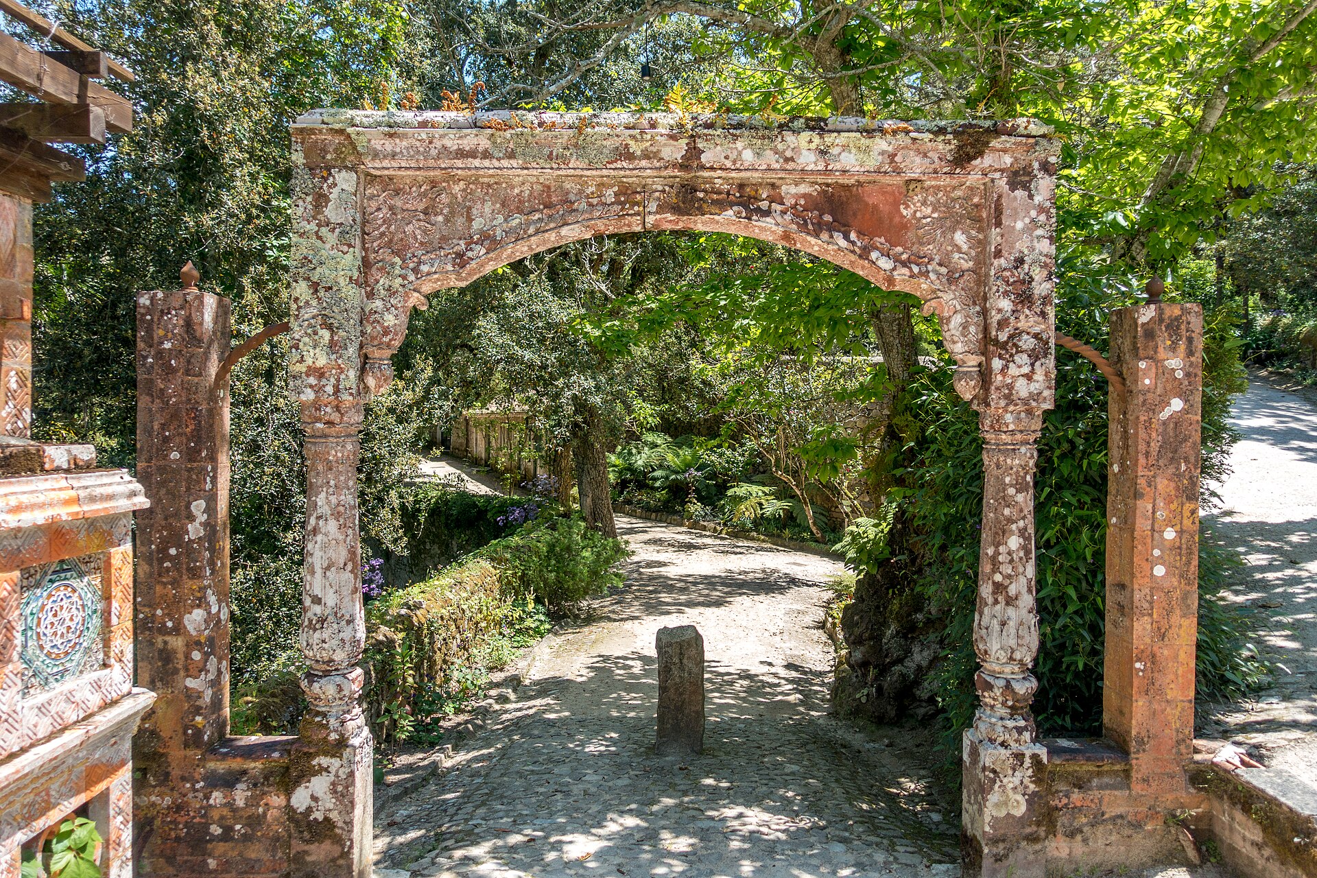 A weathered carved stone archway on a shaded gravel path in the gardens of Monserrate, Sintra