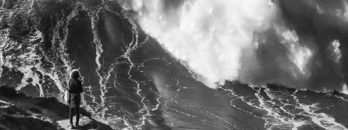 An enormous breaking wave at Praia do Norte in Nazaré with a single figure on the headland for scale