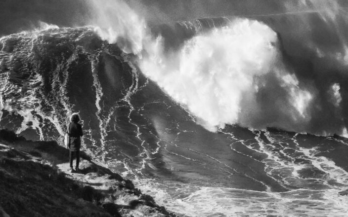 An enormous breaking wave at Praia do Norte in Nazaré with a single figure on the headland for scale