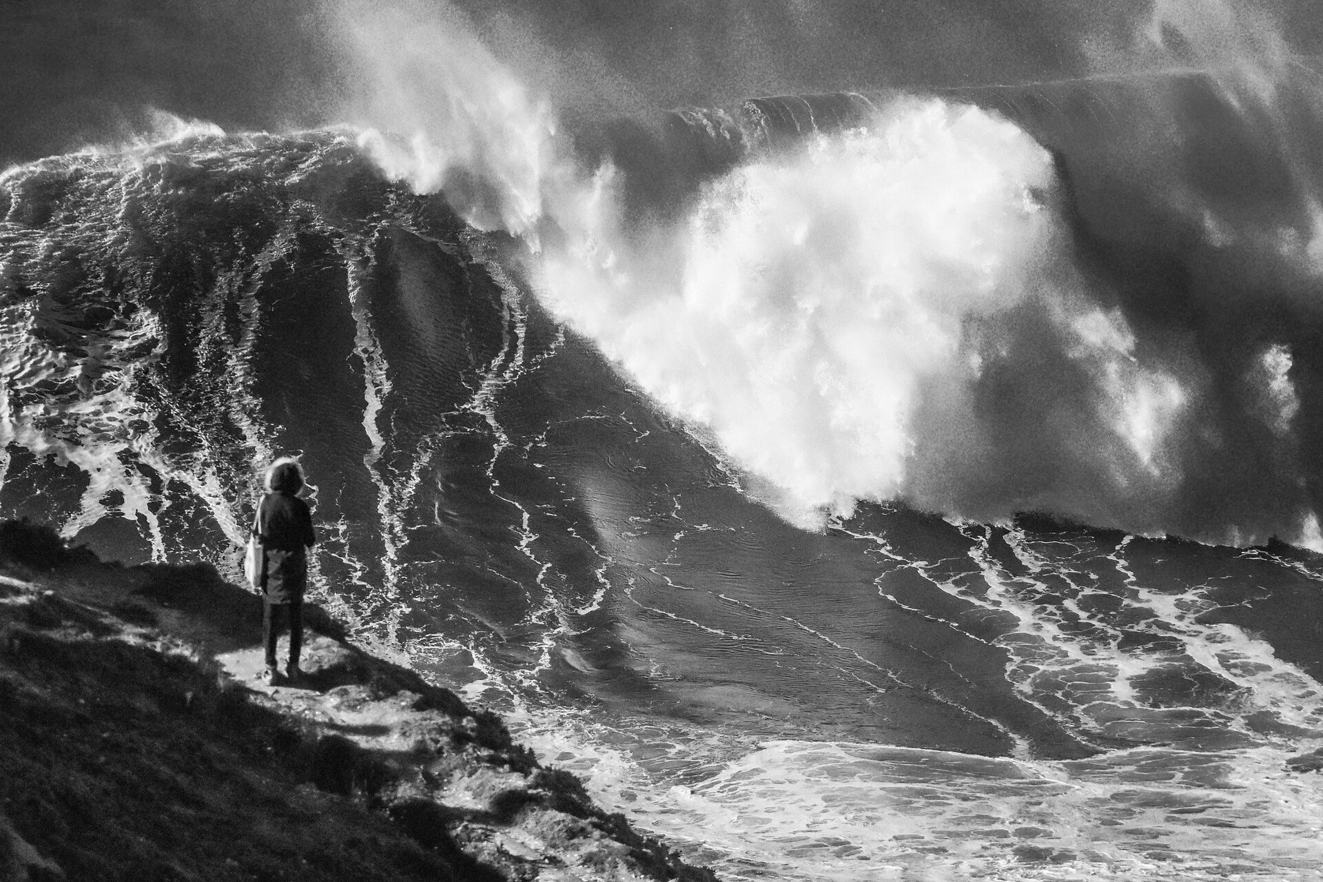 An enormous breaking wave at Praia do Norte in Nazaré with a single figure standing on the headland for scale