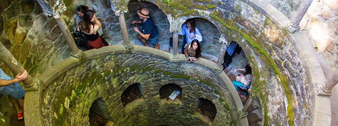 The Initiation Well at Quinta da Regaleira, a 27-metre inverted spiral staircase of mossy granite descending into the rock, with visitors on the landings