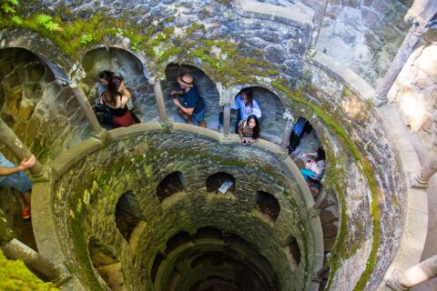 The Initiation Well at Quinta da Regaleira, a 27-metre inverted spiral staircase of mossy granite descending into the rock, with visitors on the landings