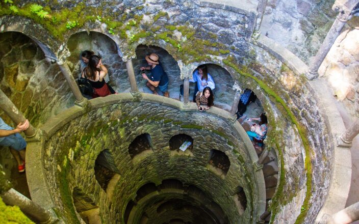 The Initiation Well at Quinta da Regaleira, a 27-metre inverted spiral staircase of mossy granite descending into the rock, with visitors on the landings