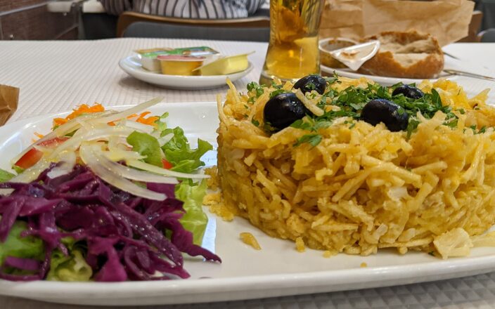 Bacalhau à Brás topped with black olives and herbs sits beside a mixed salad on a Portuguese restaurant table.