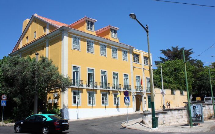 A black taxi with a green roof passes the yellow East Timorese embassy building on a sunny Lisbon street.