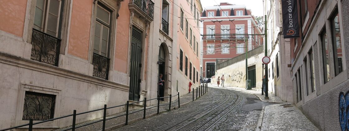 Black-and-white cobbled pavements climb Lisbon’s Calçada da Glória beside funicular rails, with pink façades and walkers farther up the hill.