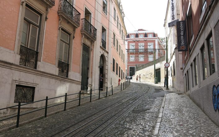 Black-and-white cobbled pavements climb Lisbon’s Calçada da Glória beside funicular rails, with pink façades and walkers farther up the hill.