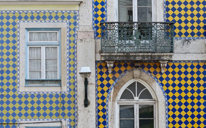 Blue and yellow azulejos surround white windows and an ornate iron balcony on a Lisbon building facade.