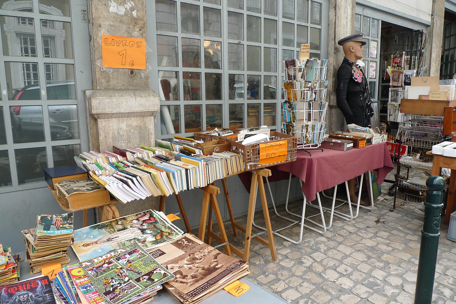 Books, magazines and postcards cover outdoor tables at Lisbon’s Feira da Ladra, beside a shop doorway and uniformed mannequin.