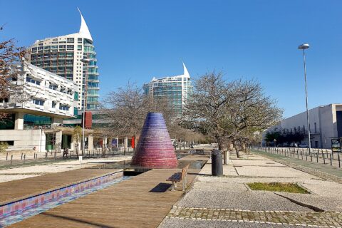 A broad promenade in Lisbon’s Parque das Nações passes a colorful conical fountain, benches, trees and modern towers.