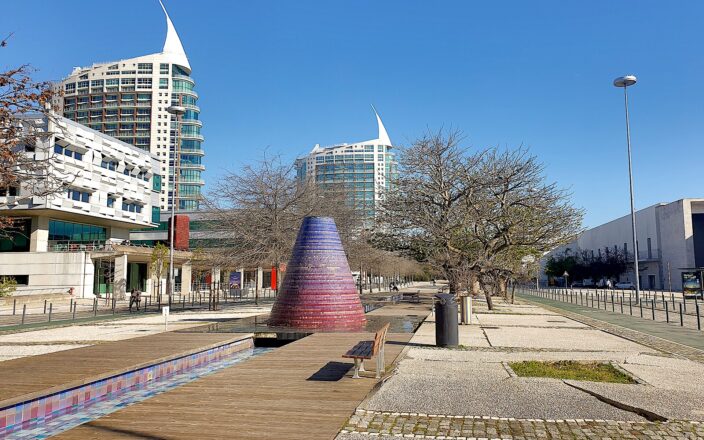 A broad promenade in Lisbon’s Parque das Nações passes a colorful conical fountain, benches, trees and modern towers.