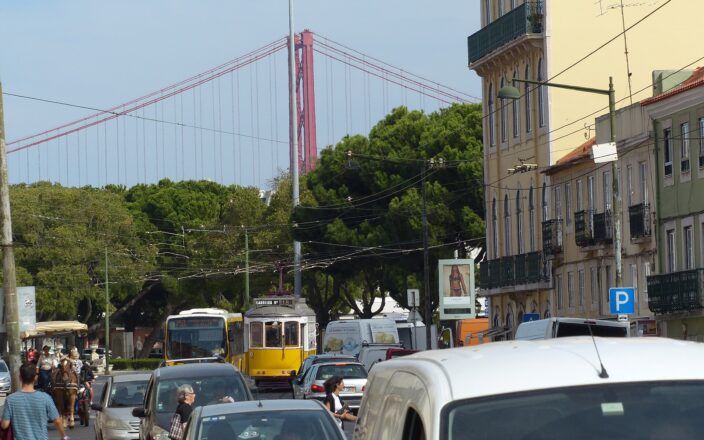 Cars and vans crowd a Lisbon street alongside a yellow tram, with the red 25 de Abril Bridge rising behind trees.