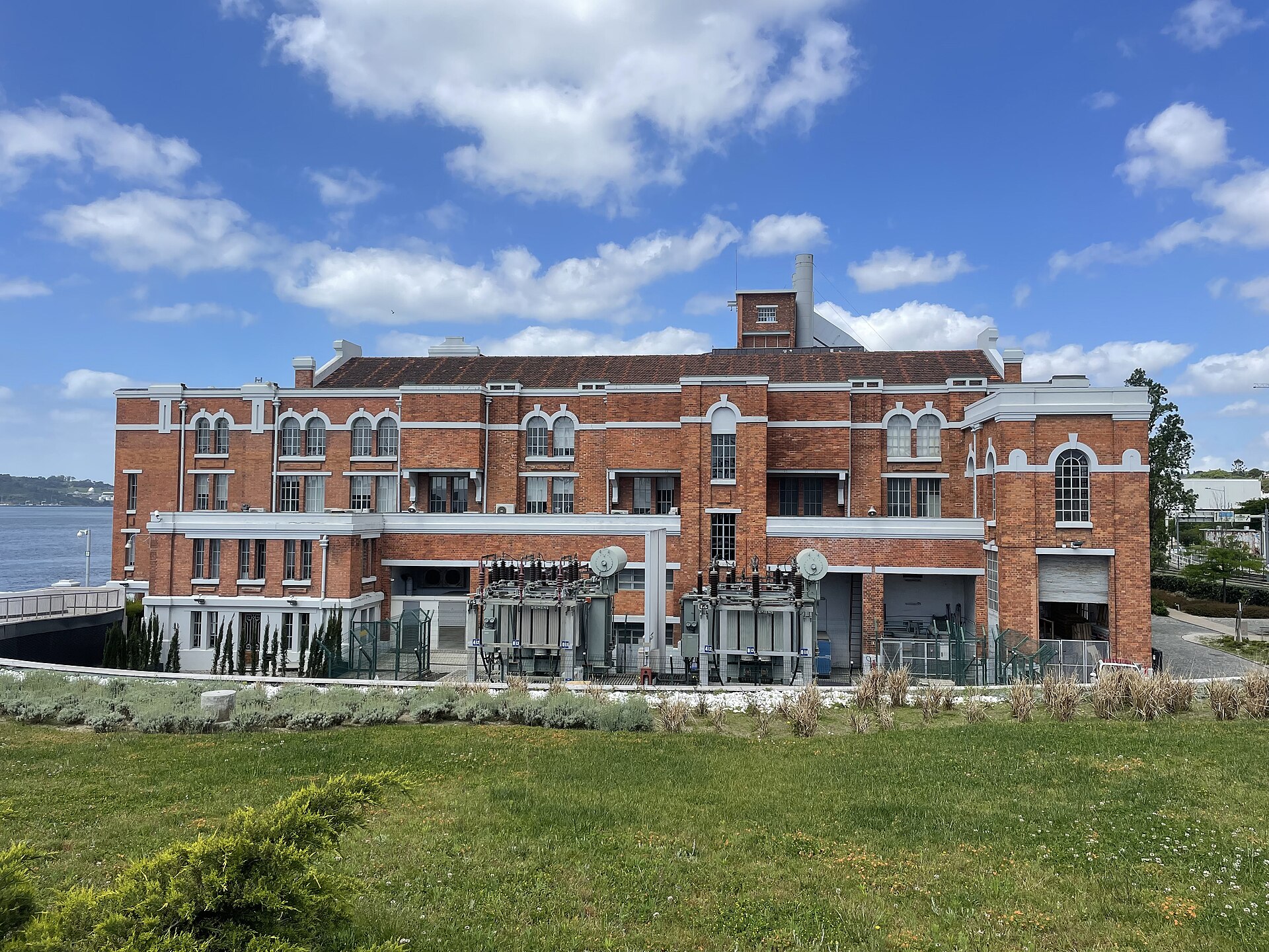 Central Tejo’s red-brick façade and electrical equipment face a grassy foreground beside the Tagus in Belém, Lisbon.