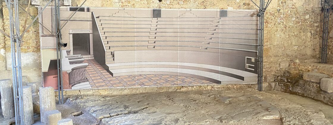 Column fragments and excavated stonework at Lisbon’s Roman theatre sit beneath a protective roof beside a reconstruction of the ancient auditorium.