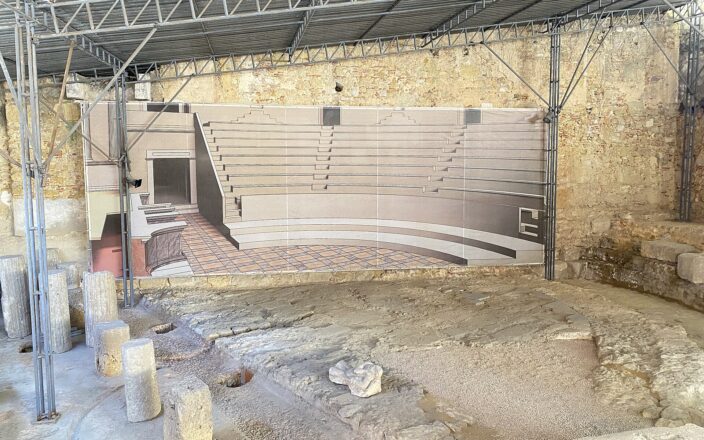 Column fragments and excavated stonework at Lisbon’s Roman theatre sit beneath a protective roof beside a reconstruction of the ancient auditorium.