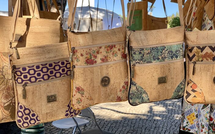 Cork shoulder bags with floral and geometric panels hang in a Lisbon retail display above patterned stone paving.