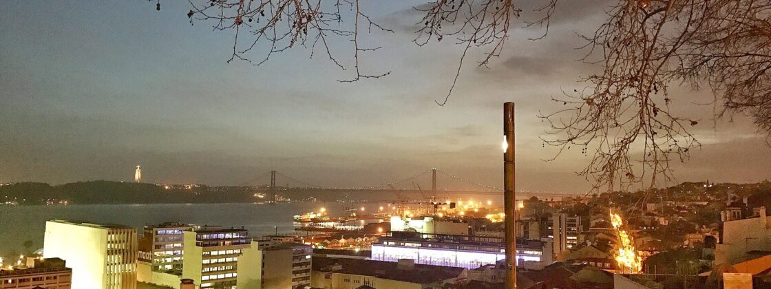 A couple sits close together among visitors at Lisbon's Santa Catarina viewpoint, overlooking the Tagus and illuminated 25 de Abril Bridge at dusk.