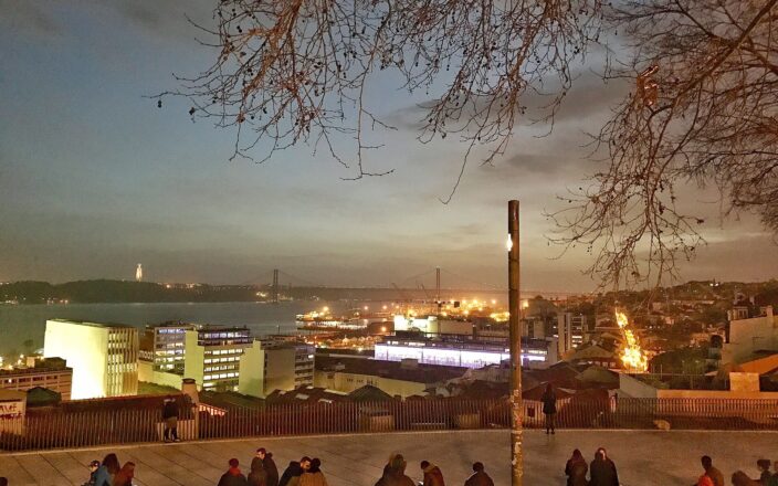 A couple sits close together among visitors at Lisbon's Santa Catarina viewpoint, overlooking the Tagus and illuminated 25 de Abril Bridge at dusk.