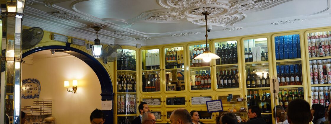 Customers gather at the counter of Pastéis de Belém in Lisbon beneath an ornate ceiling and yellow cabinets filled with bottles.