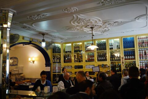 Customers gather at the counter of Pastéis de Belém in Lisbon beneath an ornate ceiling and yellow cabinets filled with bottles.