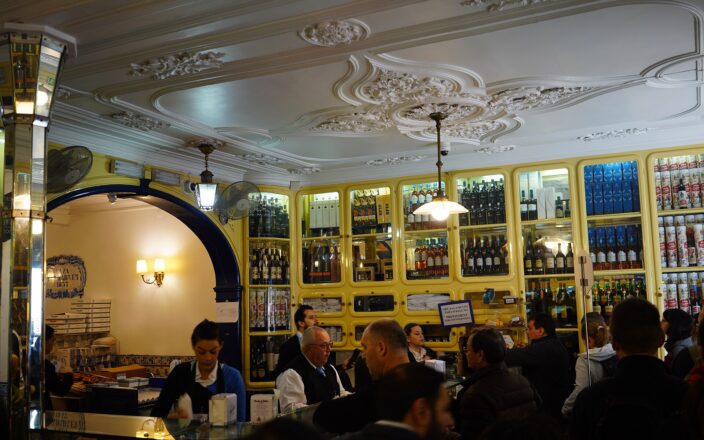 Customers gather at the counter of Pastéis de Belém in Lisbon beneath an ornate ceiling and yellow cabinets filled with bottles.