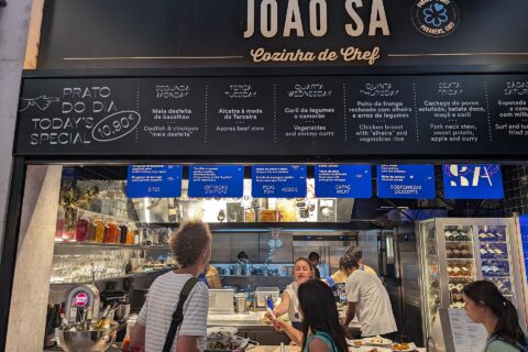 Customers stand beneath João Sá’s menu at Lisbon’s Time Out Market, where a prato do dia board advertises a €10.90 special.