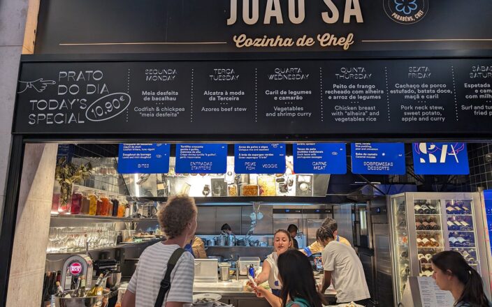 Customers stand beneath João Sá’s menu at Lisbon’s Time Out Market, where a prato do dia board advertises a €10.90 special.
