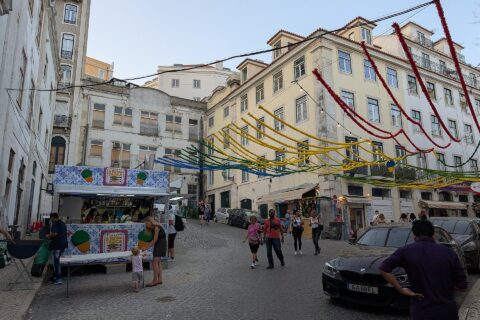 Food stalls and pedestrians fill a sloping Lisbon junction beneath colourful Santos Populares garlands stretched between apartment buildings.