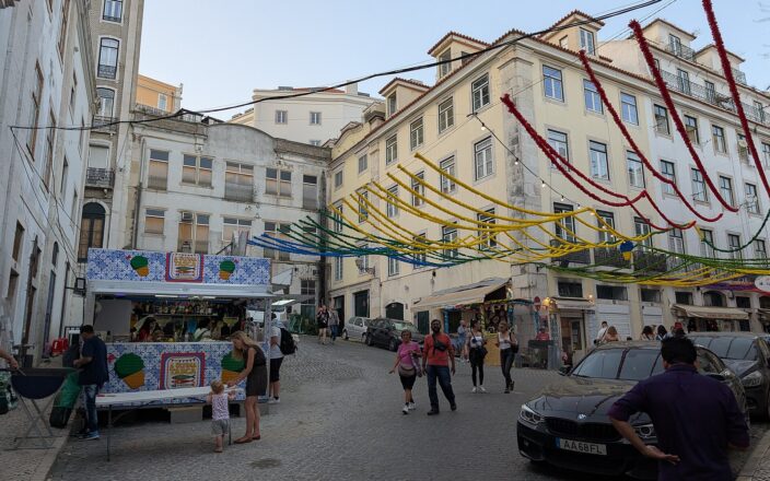 Food stalls and pedestrians fill a sloping Lisbon junction beneath colourful Santos Populares garlands stretched between apartment buildings.