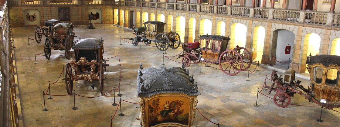 Gilded royal coaches stand beneath a painted ceiling and portrait-lined balcony in the National Coach Museum's old riding hall in Belém.