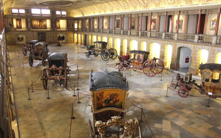 Gilded royal coaches stand beneath a painted ceiling and portrait-lined balcony in the National Coach Museum's old riding hall in Belém.