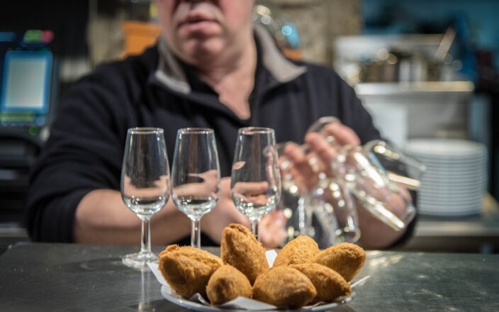 Golden cod croquettes sit beside three stemmed glasses on a Lisbon bar counter, with a server working behind them.