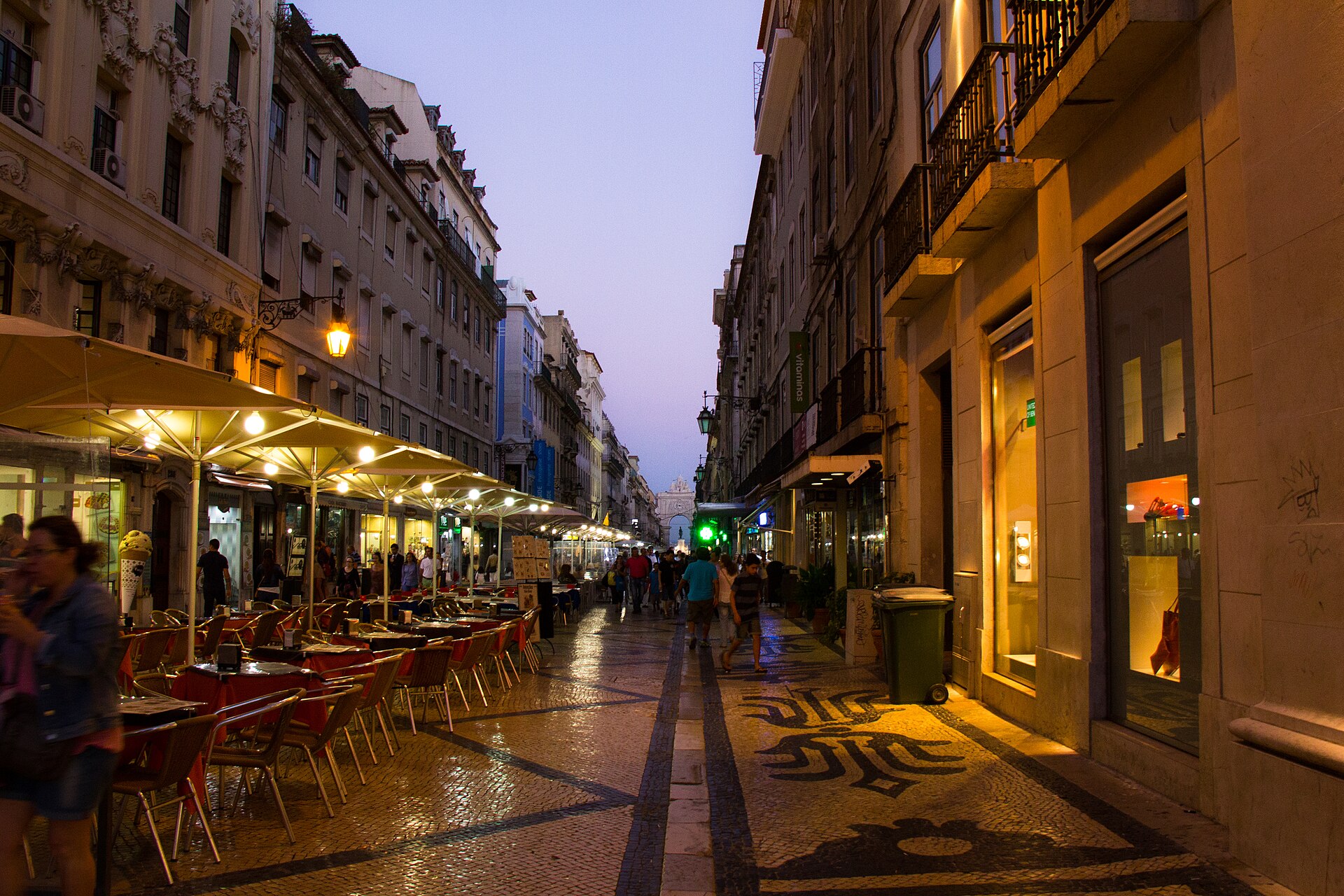 Illuminated cafe terraces and shop windows flank Lisbon’s Rua Augusta at dusk, with pedestrians walking toward the arch.