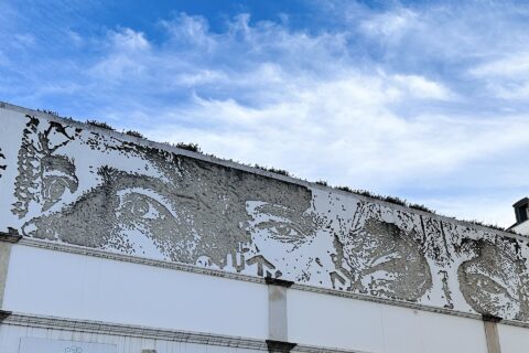 Large faces carved into a long white wall in Lisbon reveal rough grey plaster beneath a blue, cloud-streaked sky.