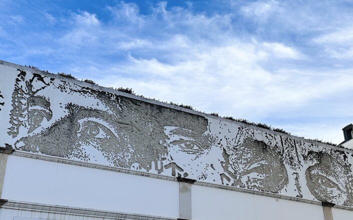 Large faces carved into a long white wall in Lisbon reveal rough grey plaster beneath a blue, cloud-streaked sky.
