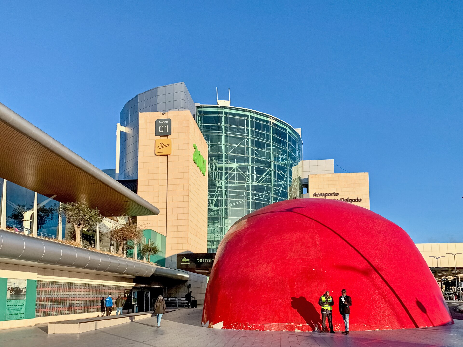 Lisbon Airport Terminal 1 rises behind the red dome of Aeroporto metro station as pedestrians cross the forecourt.