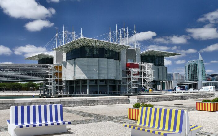 Lisbon Oceanário rises above the water behind colorful striped benches, with white roof masts and exposed external staircases.