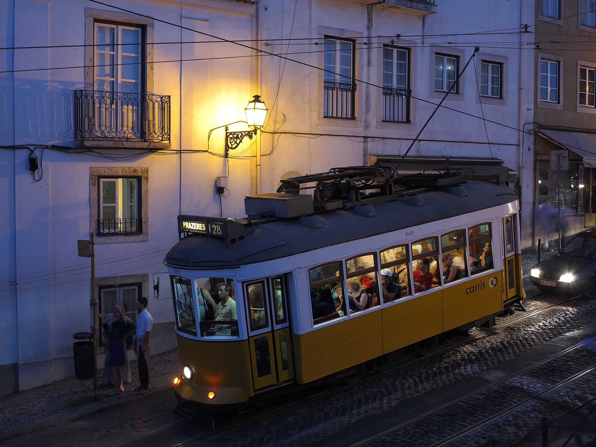 Lisbon Tram 28 carries passengers along a cobbled Alfama street at dusk beneath a glowing wall lamp.
