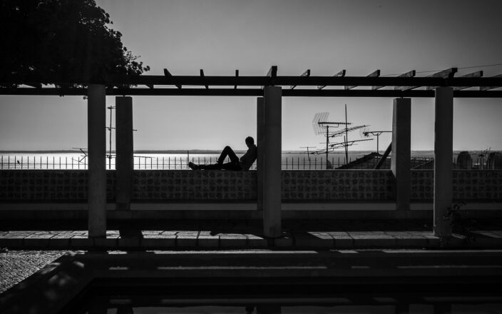 A lone visitor sits beneath the pergola at Lisbon’s Miradouro de Santa Luzia, silhouetted against the Tagus River.