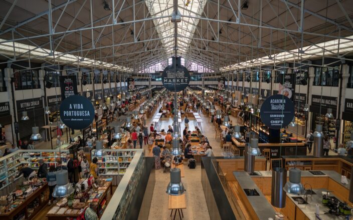 Long communal tables and food stalls fill Lisbon’s Time Out Market beneath iron roof trusses and a large circular sign.