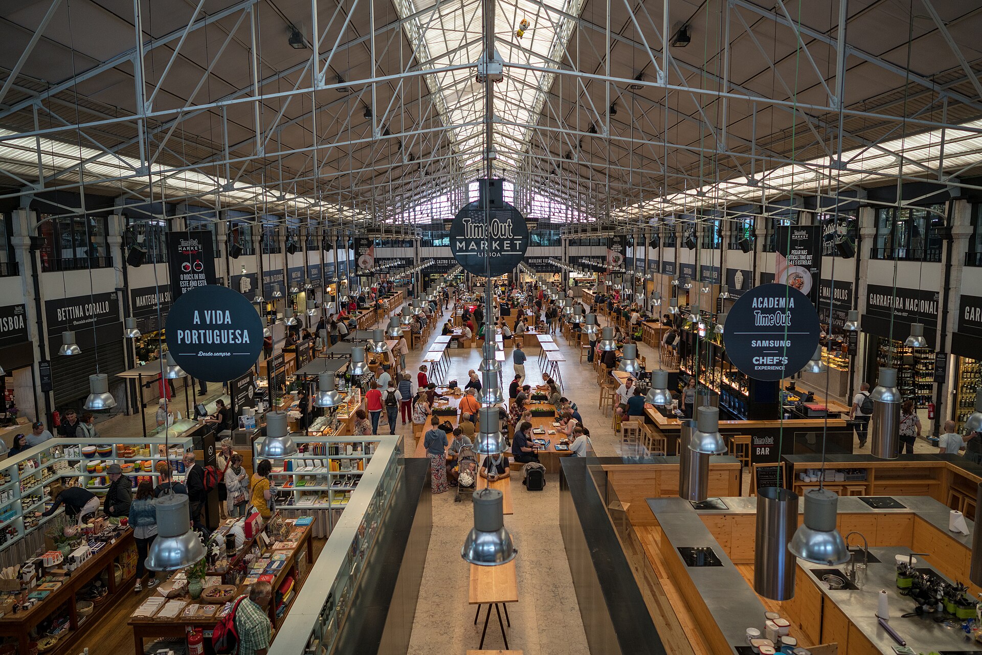 Long communal tables and food stalls fill Lisbon’s Time Out Market beneath iron roof trusses and a large circular sign.