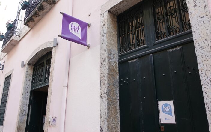 Lost Inn Lisbon Hostel has purple signs beside dark green doors in a pale pink building with stone surrounds.