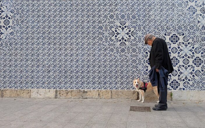 A man and his harnessed dog pause beside the blue-and-white azulejo wall at Paço da Rainha in Lisbon.