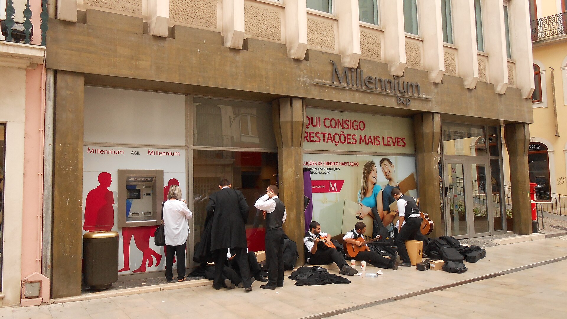 A Multibanco ATM is set into a Millennium bcp branch in Coimbra, beside students resting with guitars on the pavement.