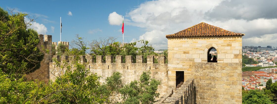 A narrow rampart leads to a stone tower at São Jorge Castle, with Lisbon rooftops visible beyond the walls.