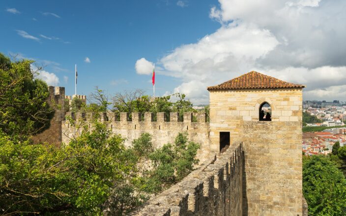 A narrow rampart leads to a stone tower at São Jorge Castle, with Lisbon rooftops visible beyond the walls.