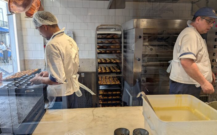 Pastry cooks prepare pastéis de nata in a Lisbon kitchen beside metal moulds, a custard tub and racks of baked tarts.