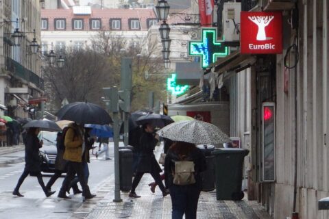 Pedestrians carry umbrellas across a rain-soaked Lisbon street beside shopfronts, glowing pharmacy crosses and traditional street lamps.